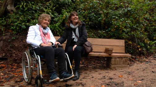 A woman sat on a bench with another woman in a wheelchair with trees behind them and autumnal leaves scattered on the path